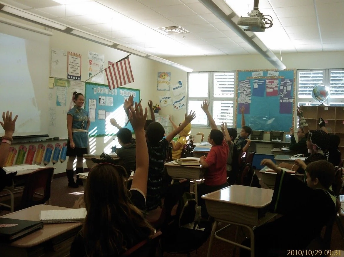 One of Ms. Lo's first Halloweens as a teacher: Elementary school classroom with students raising hands while teacher stands at front. Classroom has educational posters, American flag, globe, and sunlight coming through windows.