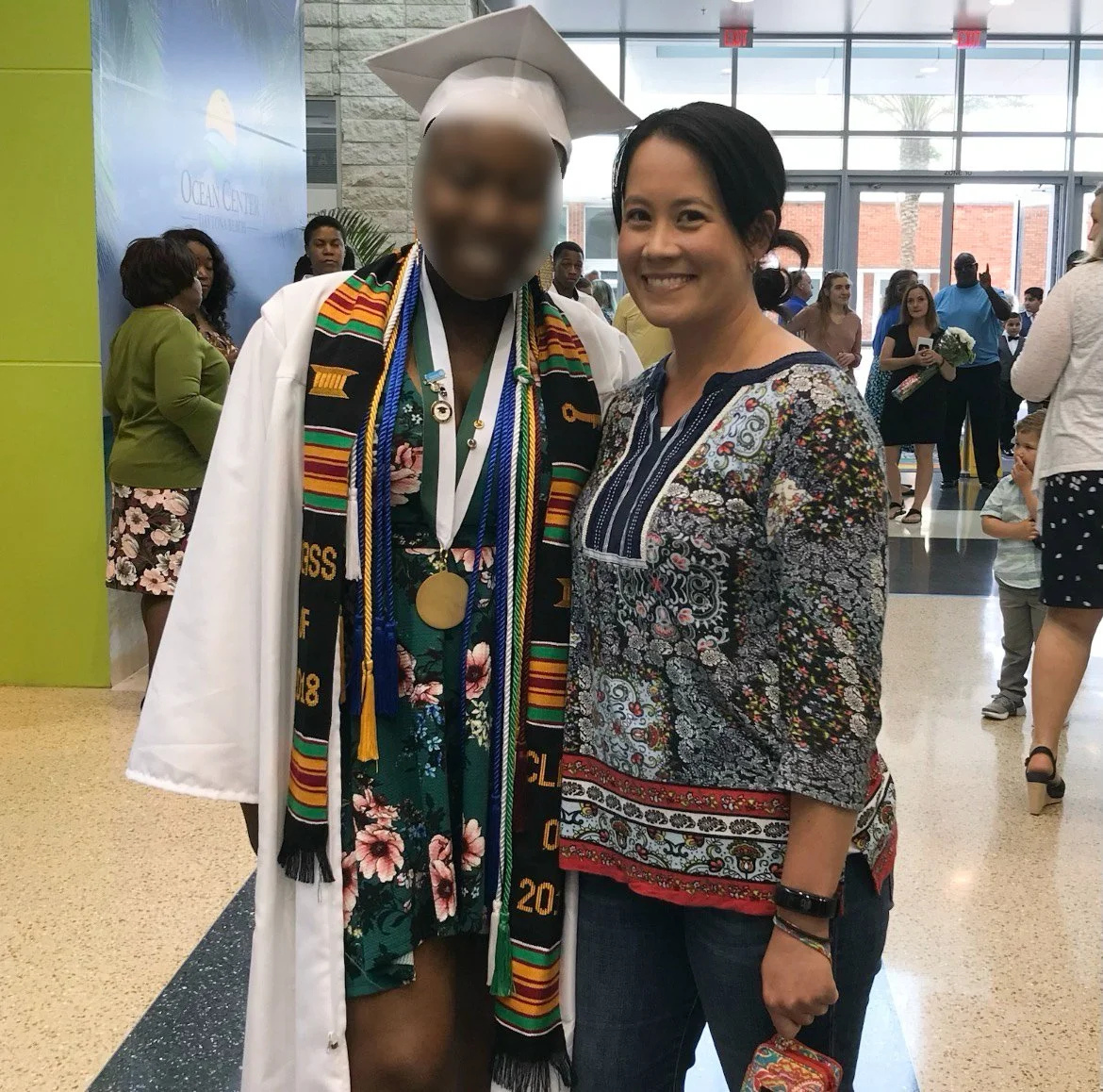 Ms. Lo with a former student at her high school graduation in Flagler County: Woman in a colorful blouse standing next to a high school graduate in academic regalia at a graduation ceremony inside a school building.