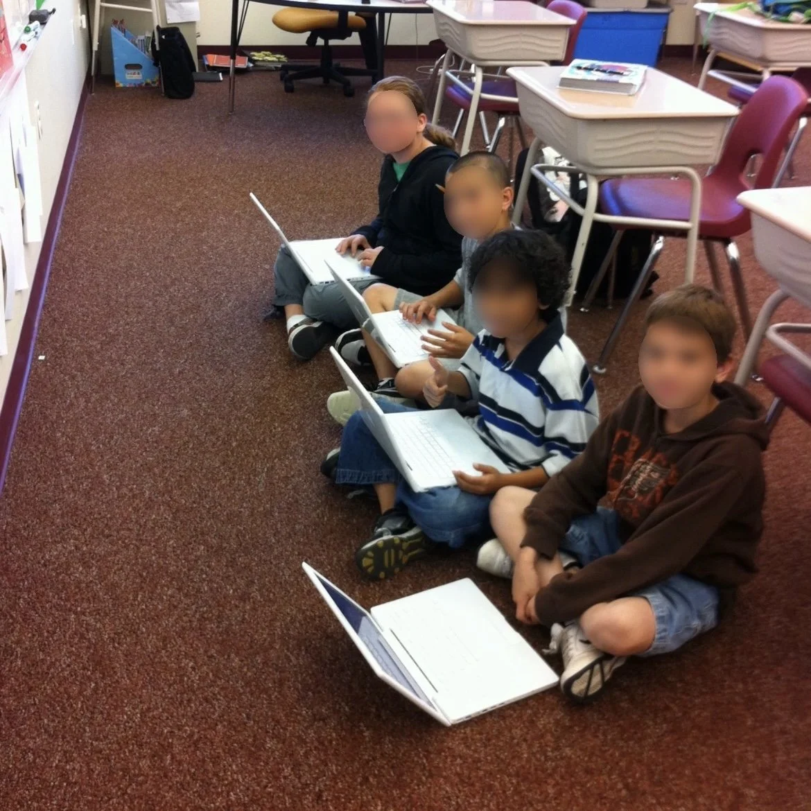 Four children sitting on the classroom floor with laptops in front of them, looking up at the camera in a classroom setting with desks and chairs in the background.
