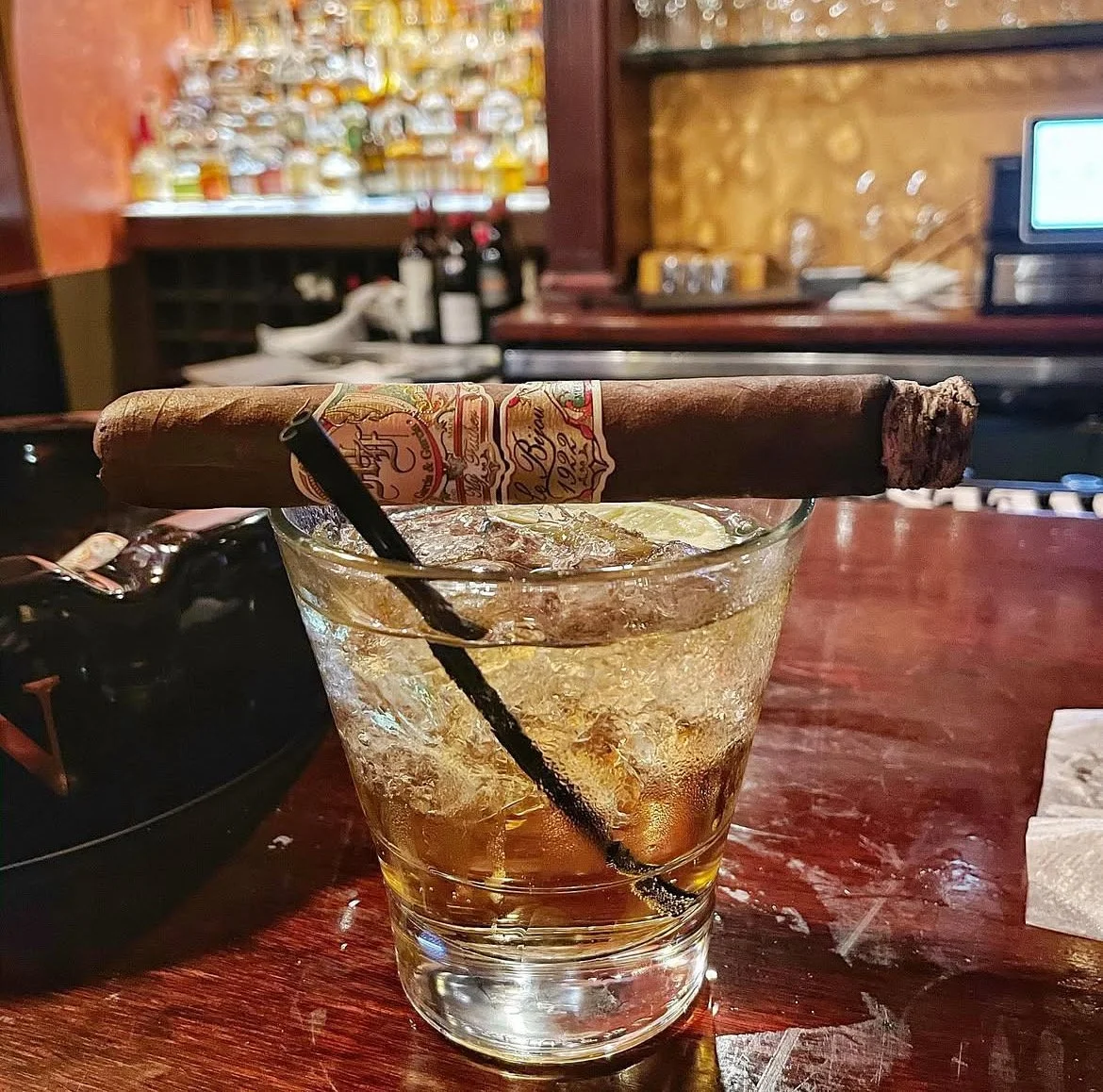 A glass of whiskey with ice and a lit cigar resting on the rim, on a wooden bar counter.
