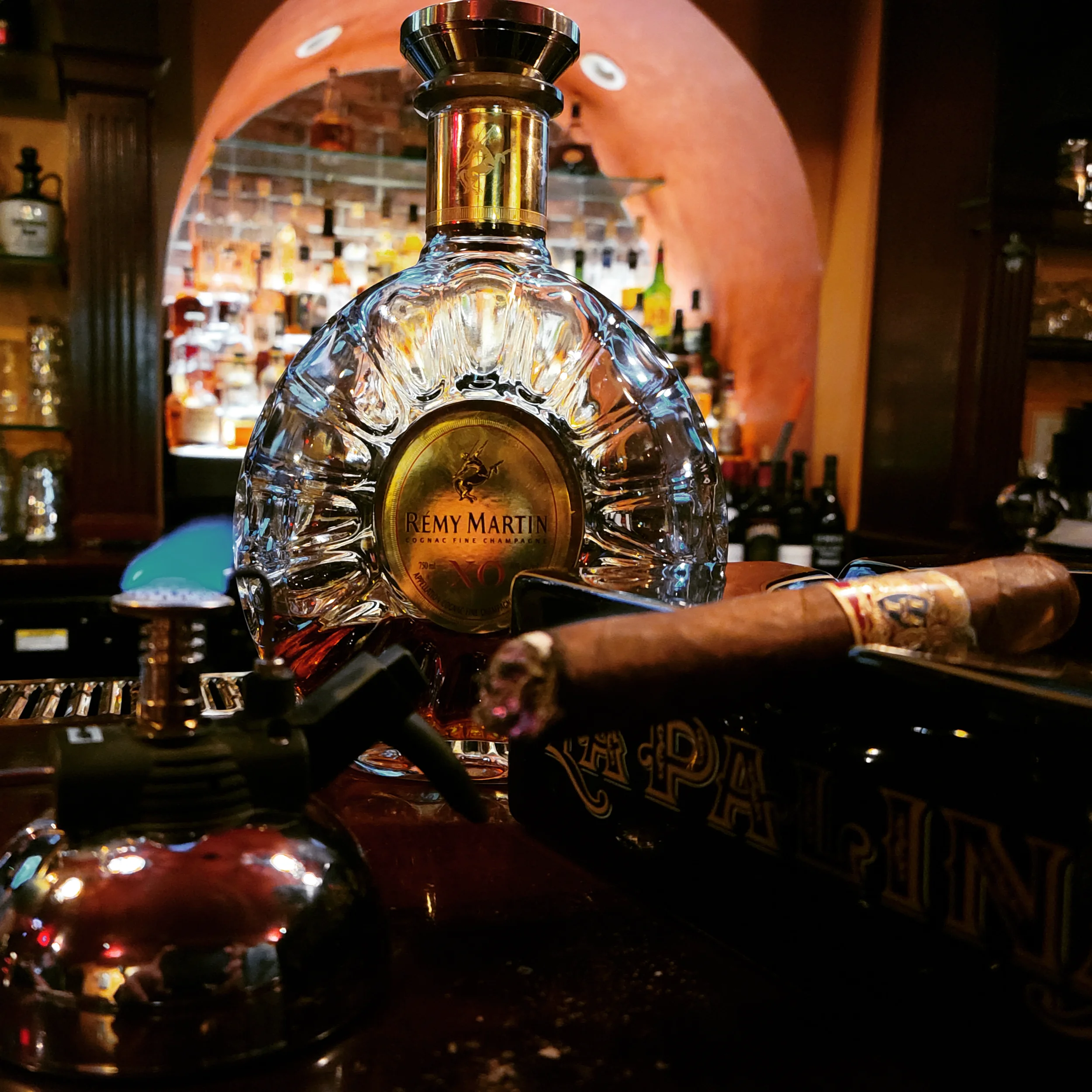 A bottle of Rémy Martin Louis XIII cognac on a bar counter, with a cigar, a black lighter, and various bottles and bar tools in the background.