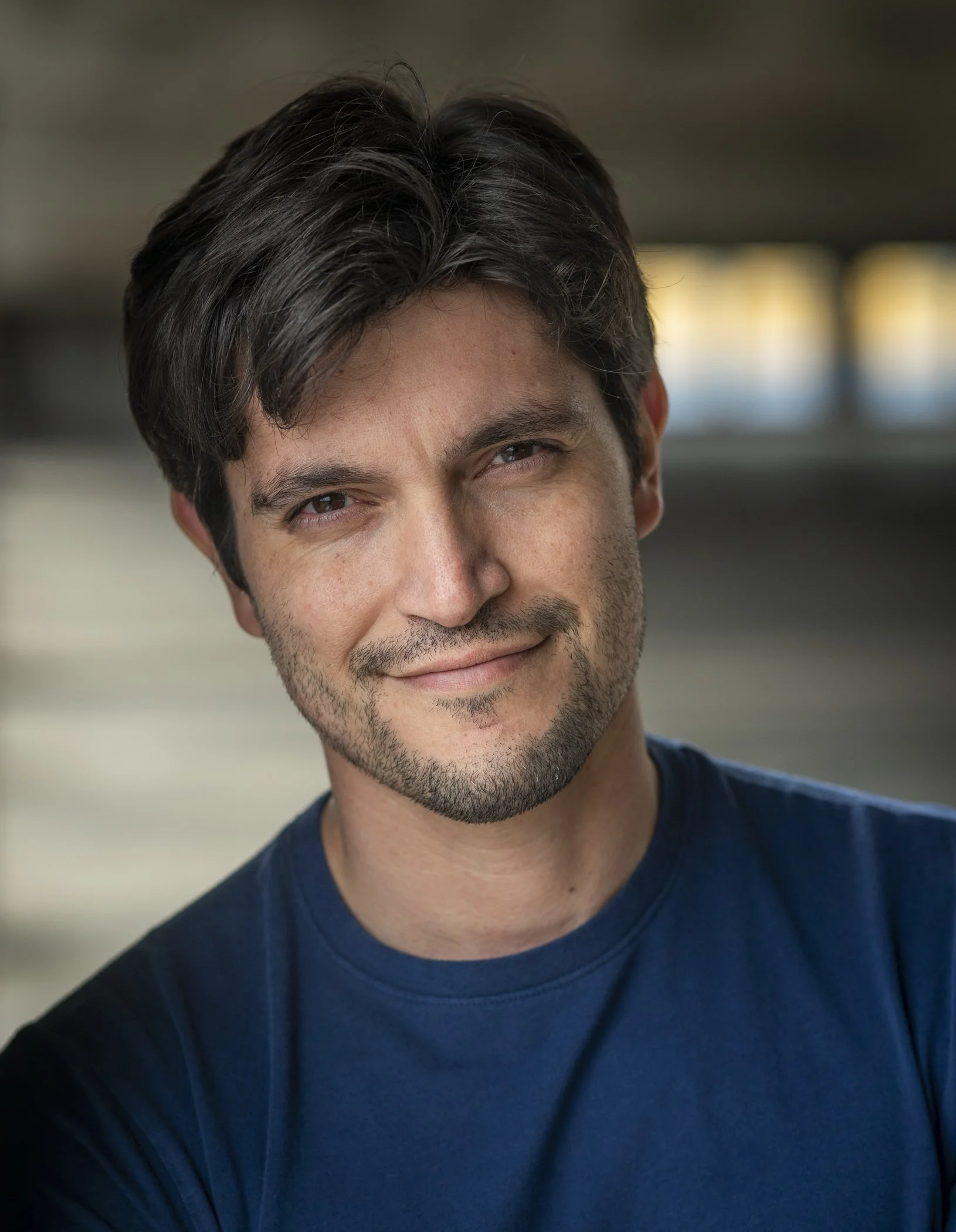 Headshot of a handsome young man with dark hair, slight beard, and a smiling expression, wearing a blue shirt.