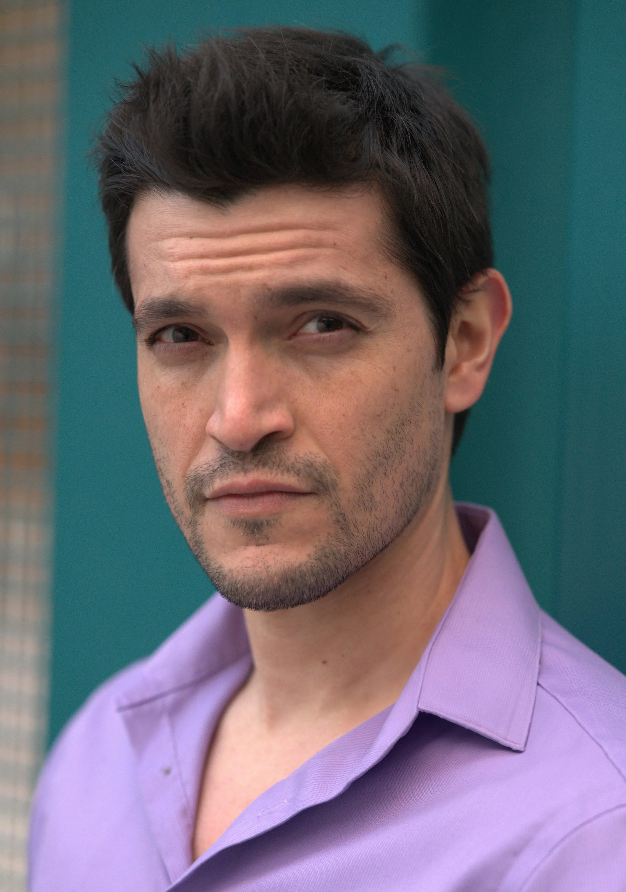 A man with dark hair and light stubble in a lavender collared shirt, standing outdoors against a teal background with a neutral expression.