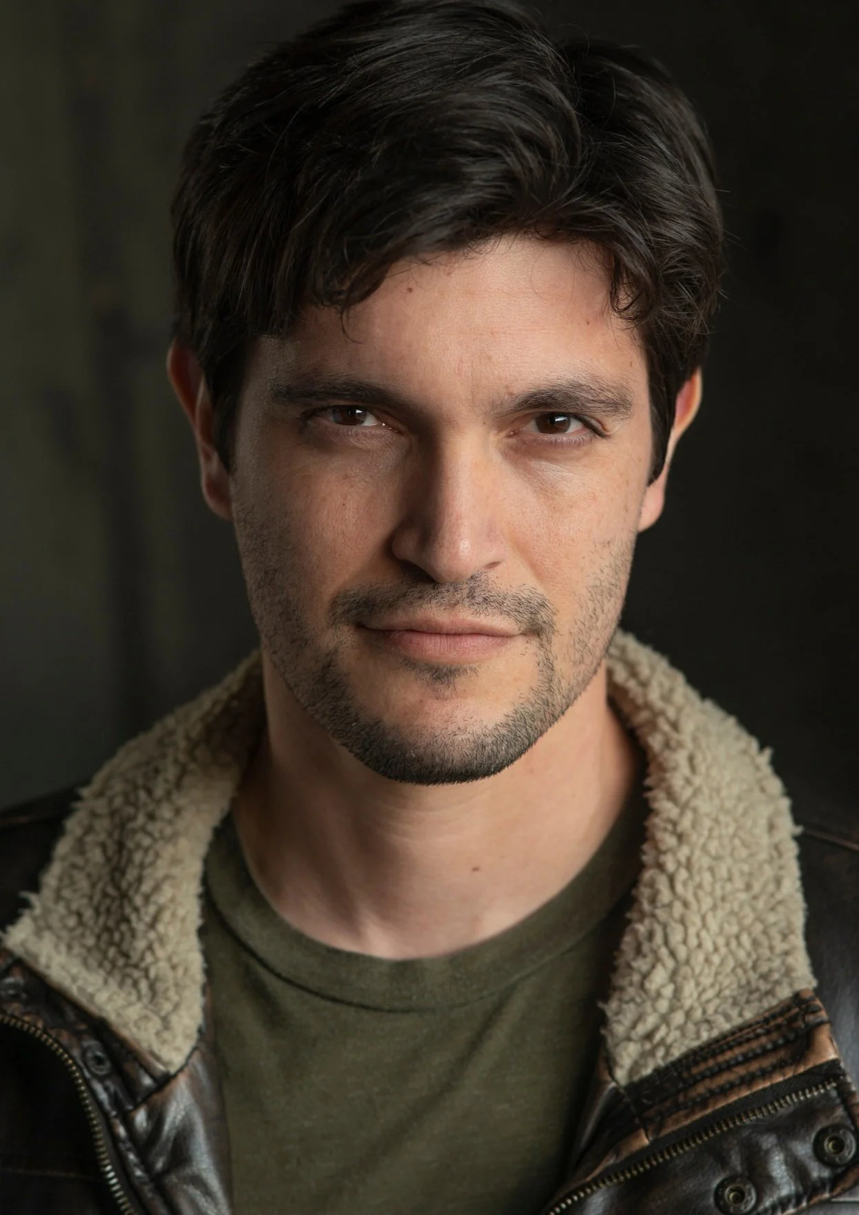 Close-up portrait of a man with dark hair, slight beard, wearing a leather jacket with a shearling collar and an olive green t-shirt, looking directly at the camera.