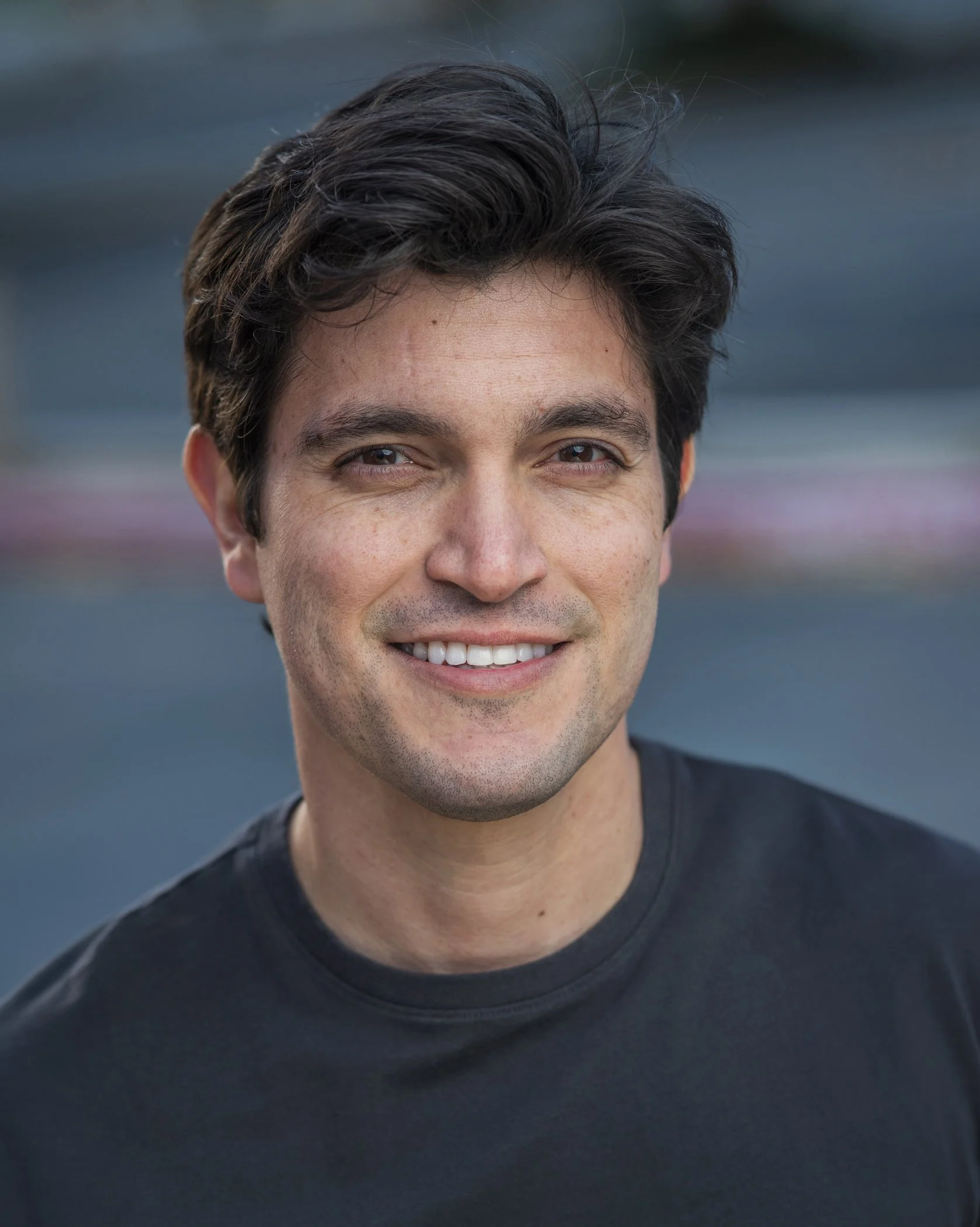 Headshot of a handsome young man with dark hair, smiling, wearing a black shirt, outdoors with blurred background.