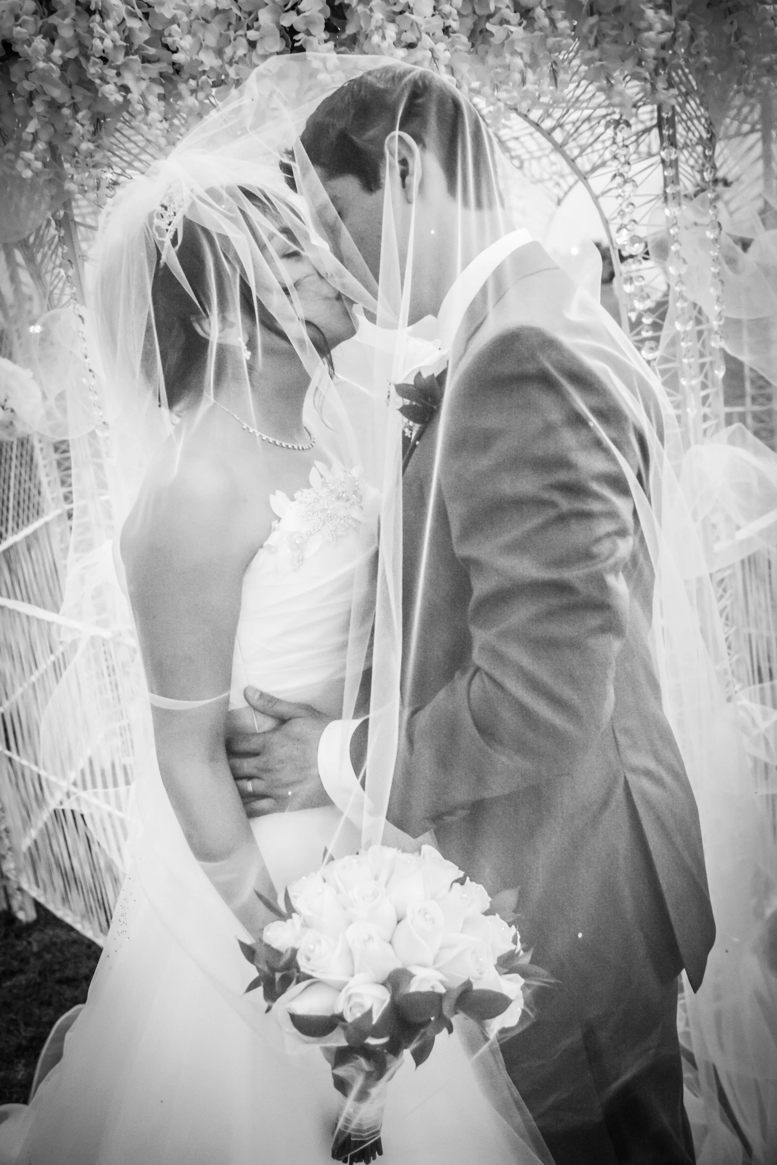 Beautiful dreamy black and white photograph of a bride and groom sharing a kiss at their wedding, with the bride holding a bouquet of roses, both partially obscured by the wedding veil Humberto Castro and Lorraine Sanchez.