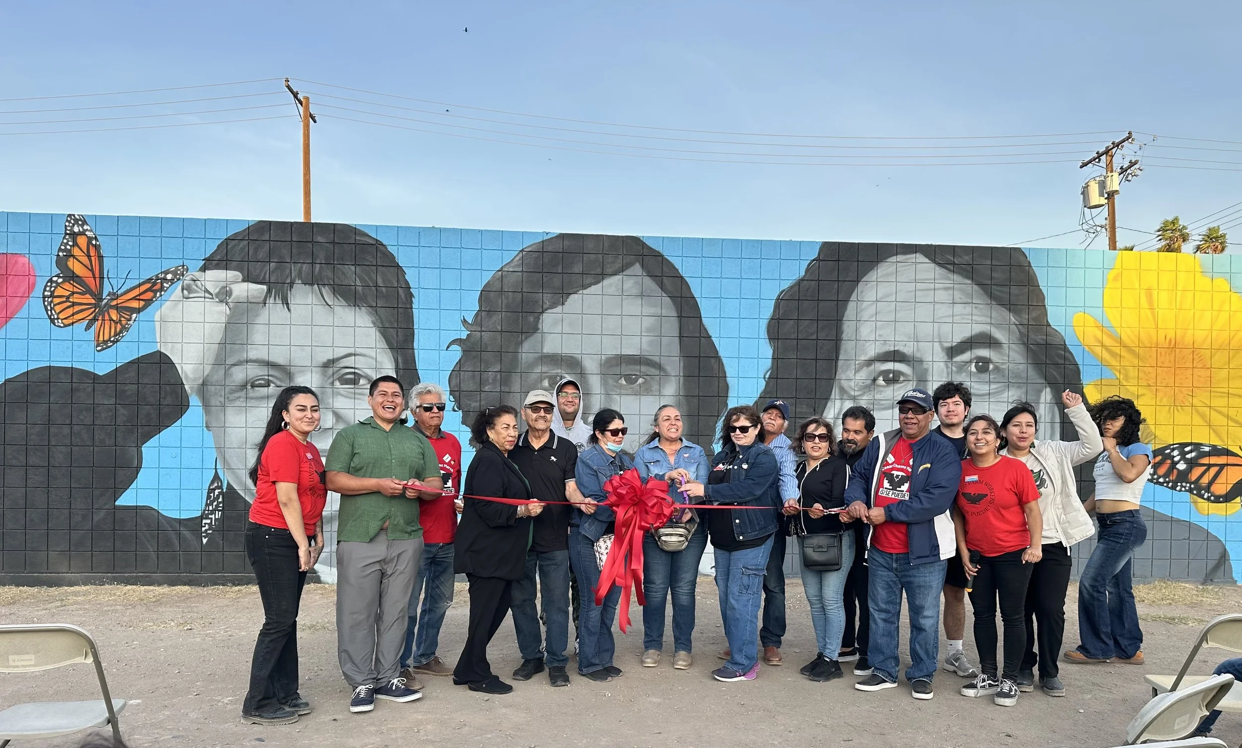 A group of folks gathered in front of a mural depicting 3 Latinx leaders in the Chicano & United Farmworker Movements during a ribbon cutting