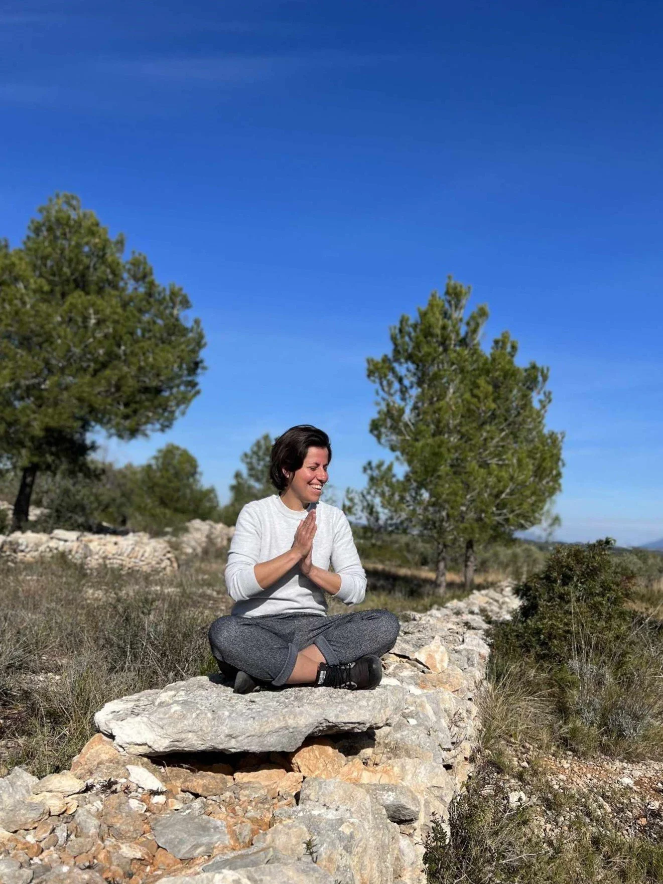 Une femme assise en position de méditation sur une pierre dans un paysage naturel avec des arbres, sous un ciel bleu.
