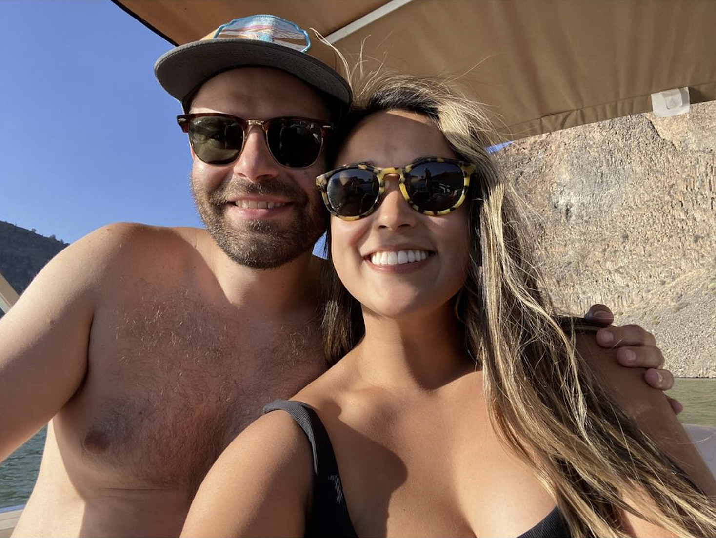 A smiling couple taking a selfie outdoors, with a rocky hillside and blue sky in the background. The man is shirtless, wearing sunglasses and a cap, while the woman is wearing sunglasses and a black top.