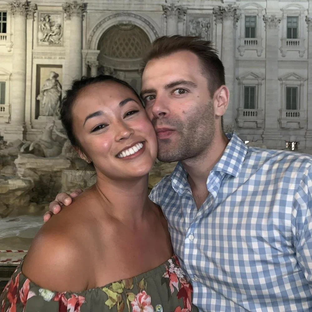 A smiling woman with dark hair and a man with short brown hair. They are outdoors with a historic building and fountain in the background.