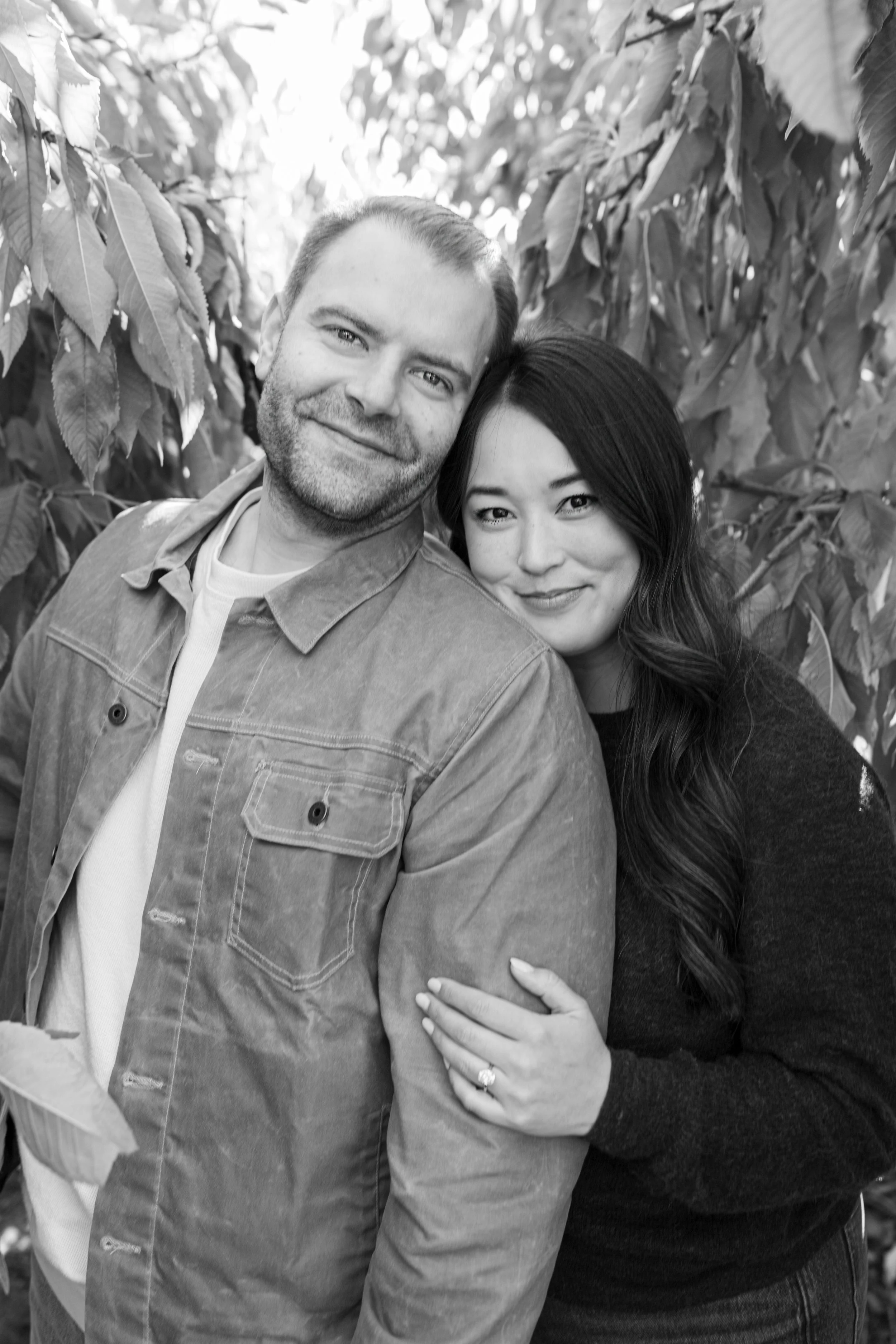 A black and white photograph of a happy couple standing close together outdoors surrounded by leafy trees. The man is smiling and has short hair and a beard, wearing a casual jacket. The woman, with long wavy hair, is smiling softly and has her hand 