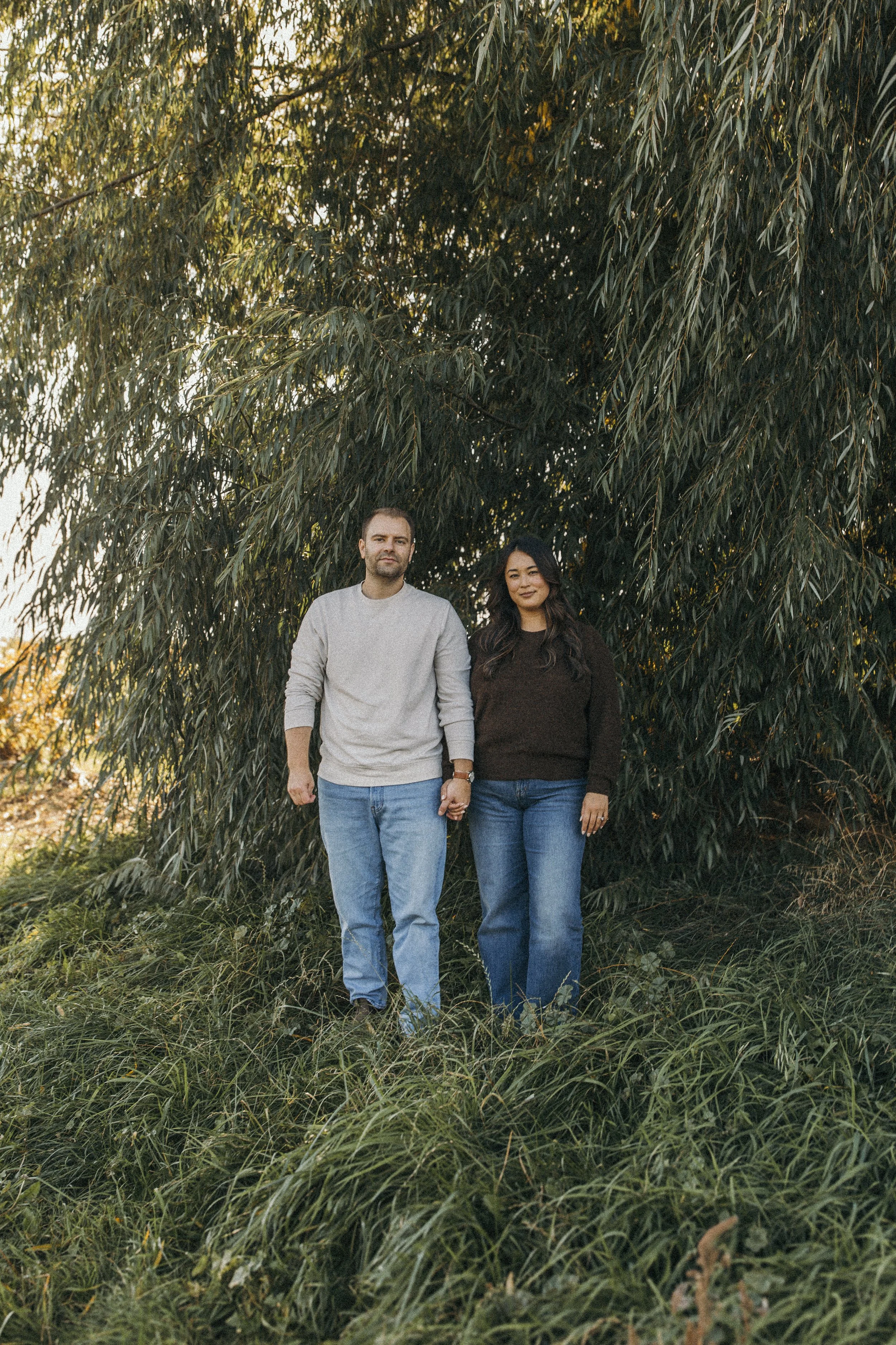A man and woman holding hands standing outdoors in front of a large, leafy tree, surrounded by grass.