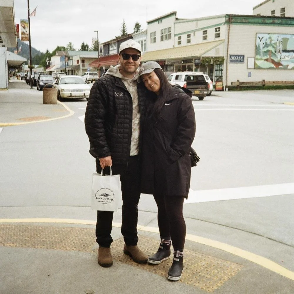 Two people, a man and a woman, standing together on a city street, smiling. The man is wearing sunglasses, a white cap, a black jacket, black pants, and brown shoes, holding a white shopping bag. The woman is wearing a gray cap, a black jacket with a