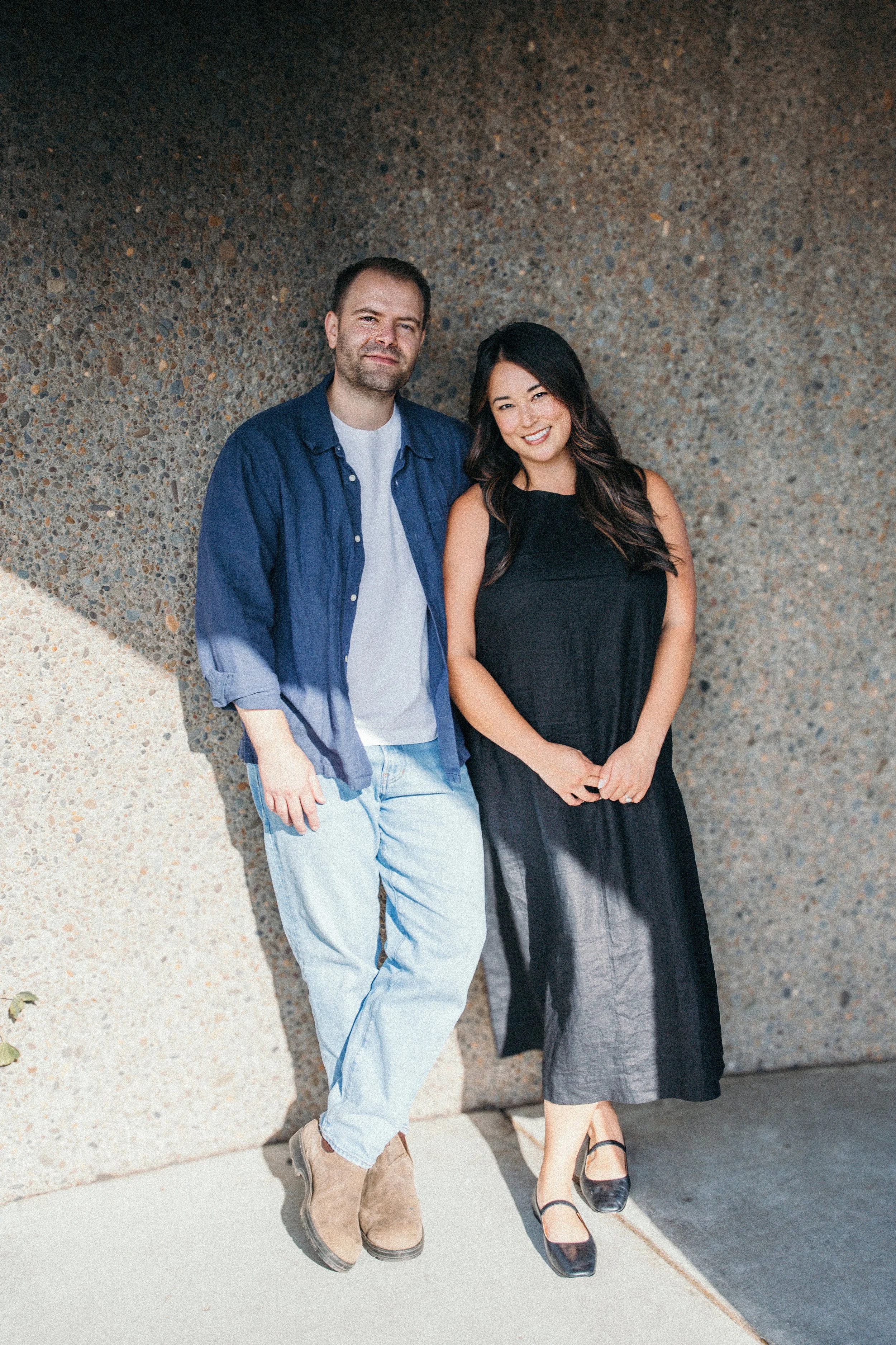 A man and woman standing together against a textured wall, smiling at the camera.