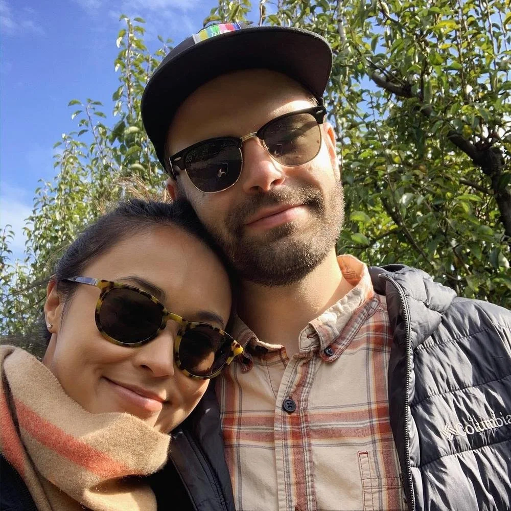 A man and woman wearing sunglasses outdoors, smiling with trees and blue sky in the background.