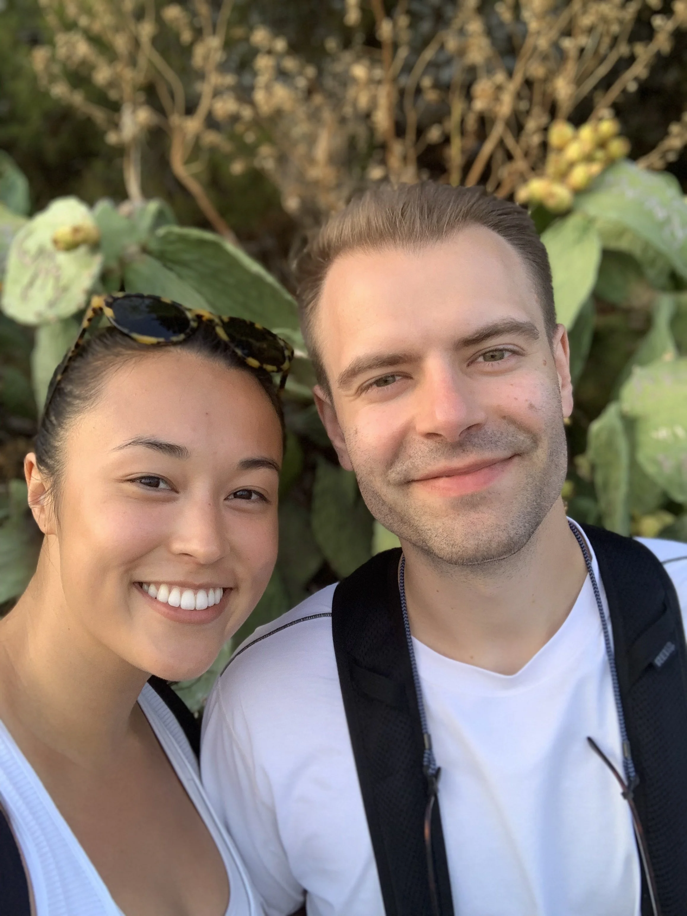 A smiling woman and man taking a selfie outdoors, with large green leafy plants in the background.