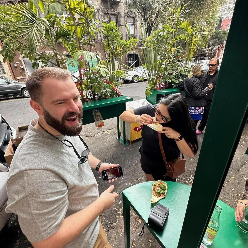 People enjoying outdoor dining with plants, a street, and parked cars in the background. Two women are eating, and a man is smiling and pointing at a table with a sandwich, a camera, and a beverage.