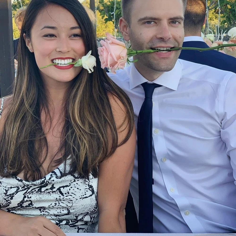 A smiling woman and man are each holding a flower in their mouths. The woman has long brown hair and is wearing a patterned top. The man has short hair, a white shirt with a dark tie, and is holding a pink flower. They are outdoors at a social event,