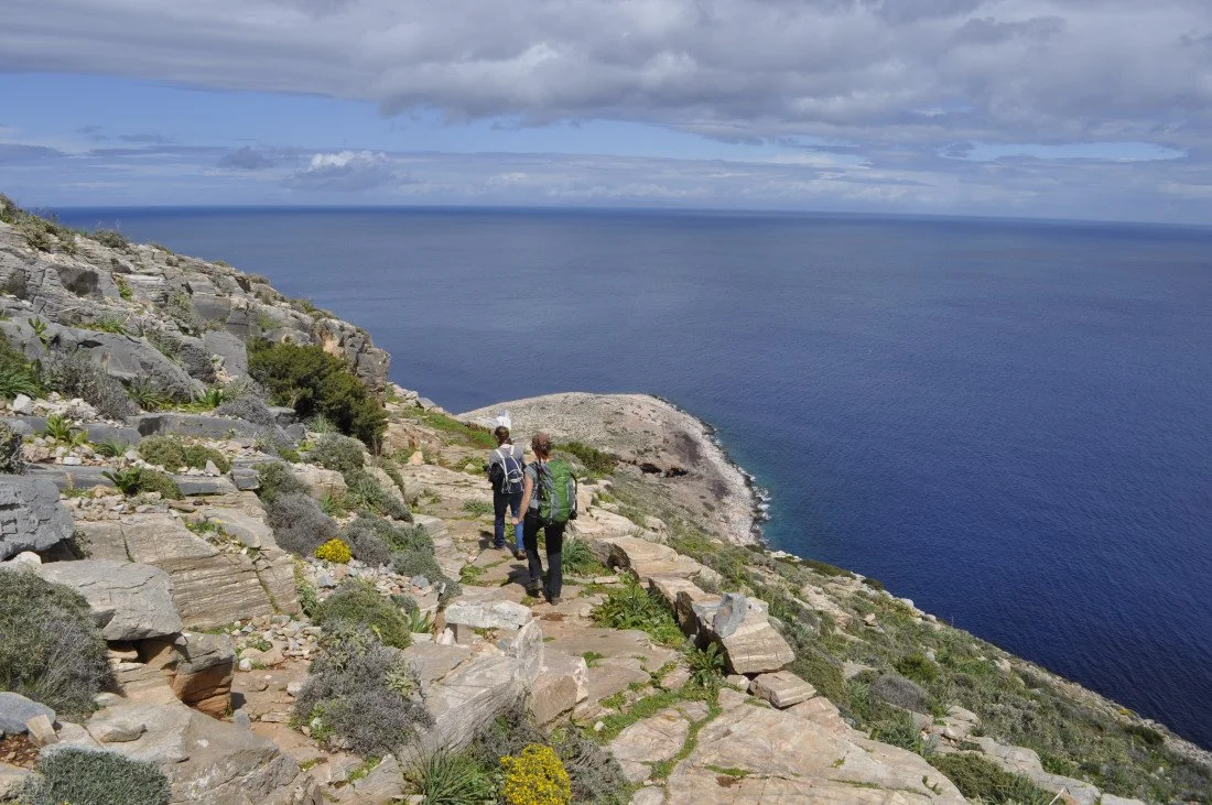 Hikers walking along a rocky coastal trail overlooking the ocean under a partly cloudy sky.