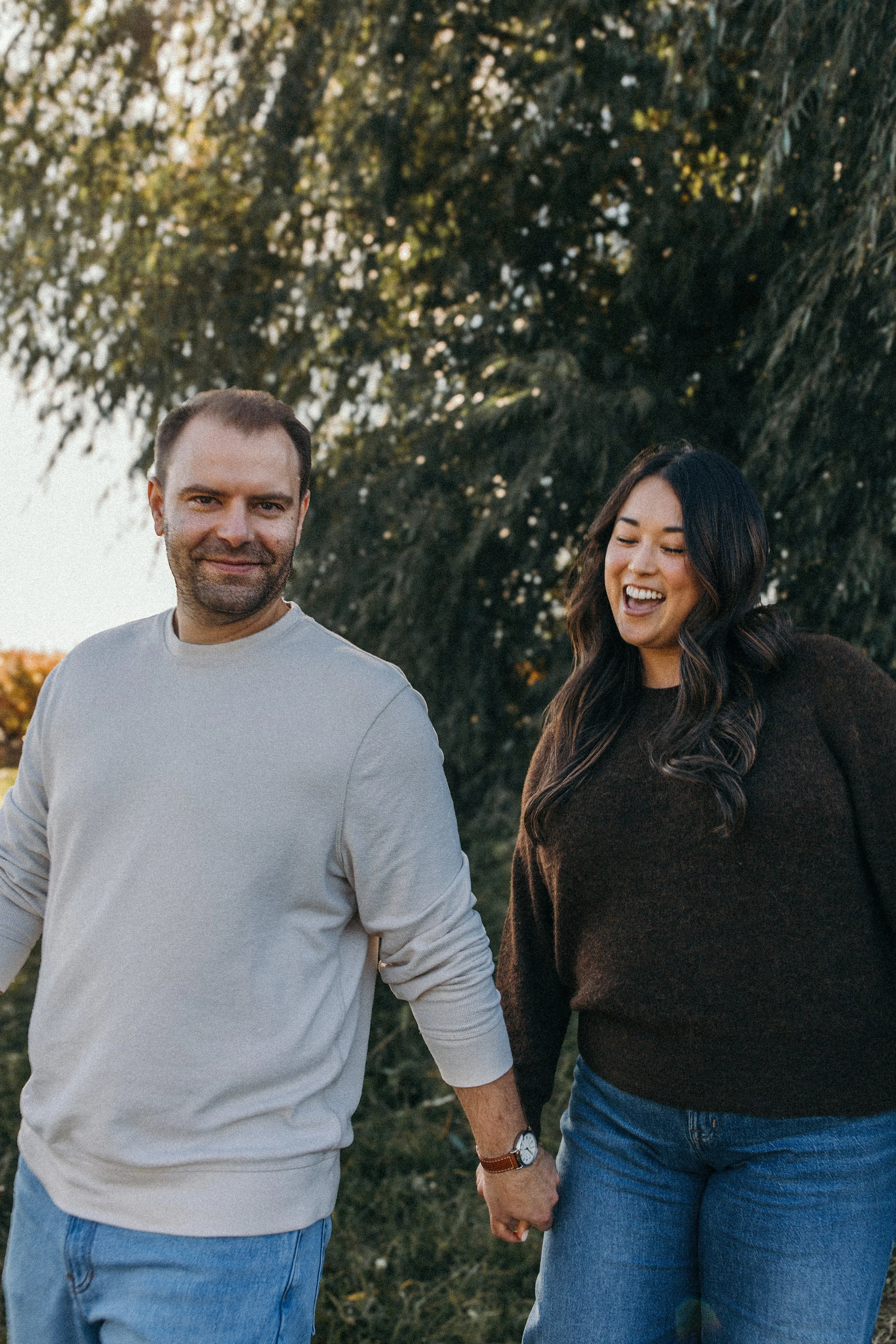 A happy couple walking outdoors, holding hands, during daytime with trees in the background.