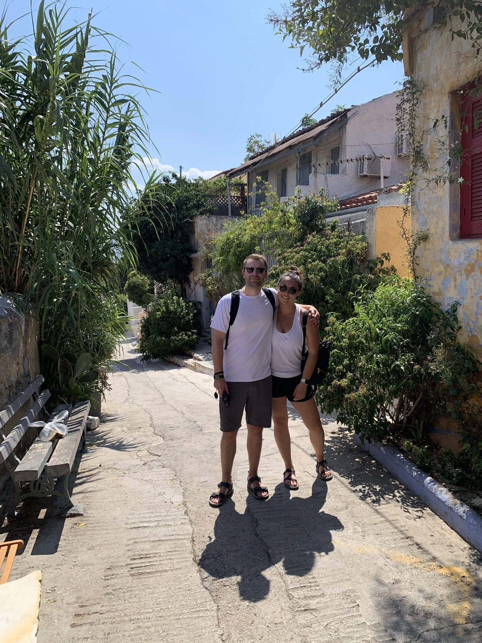 A couple standing on a sunny street with lush greenery and colorful houses, posing for a photo.