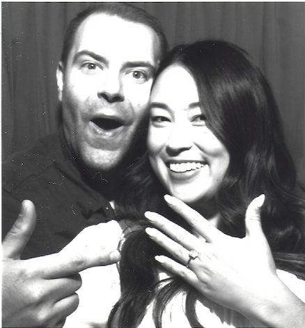 Two friends smiling and posing for a photo in front of a curtain, with one making a peace sign and the other showing off a ring on her finger.