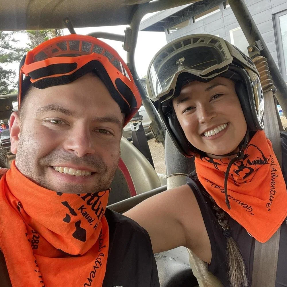 Two smiling people, wearing helmets and orange bandanas, sitting inside a rugged off-road vehicle.