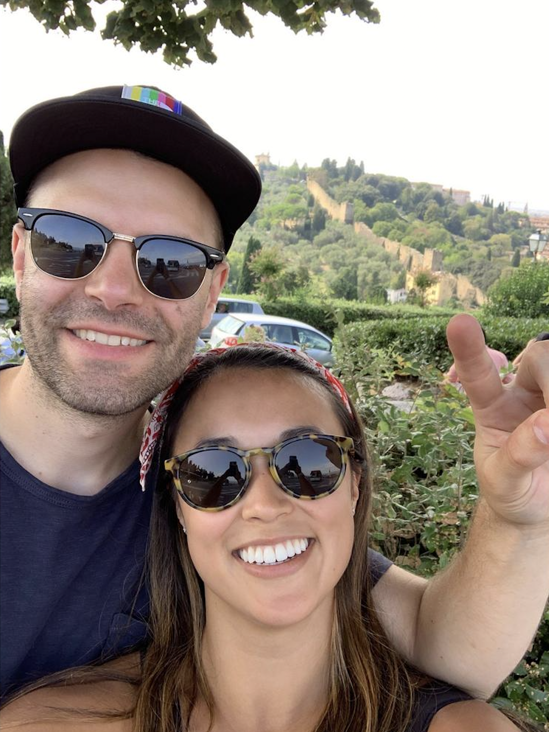 A smiling man and woman wearing sunglasses taking a selfie outdoors, with a hilly landscape and trees in the background.