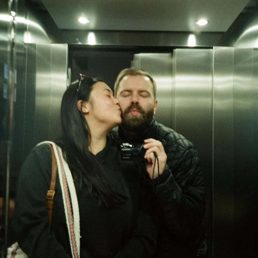 A woman giving a man a kiss on the cheek inside an elevator with reflective metal walls.