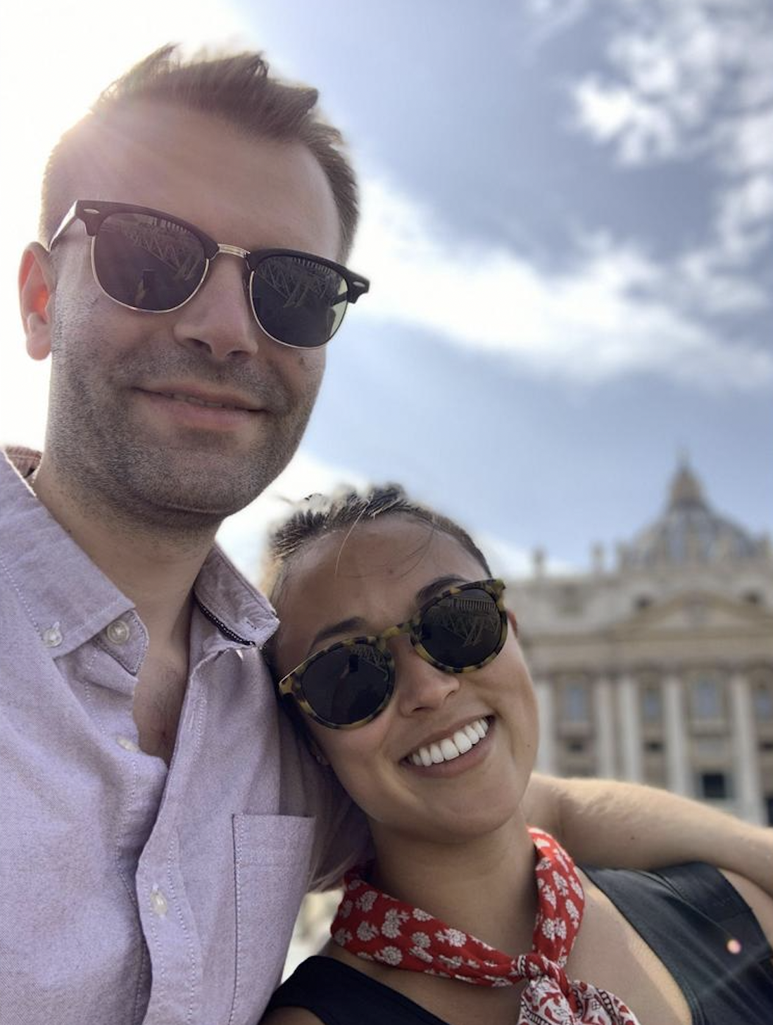 A smiling man and woman wearing sunglasses taking a selfie outdoors against a background of a historic building and blue sky with clouds.