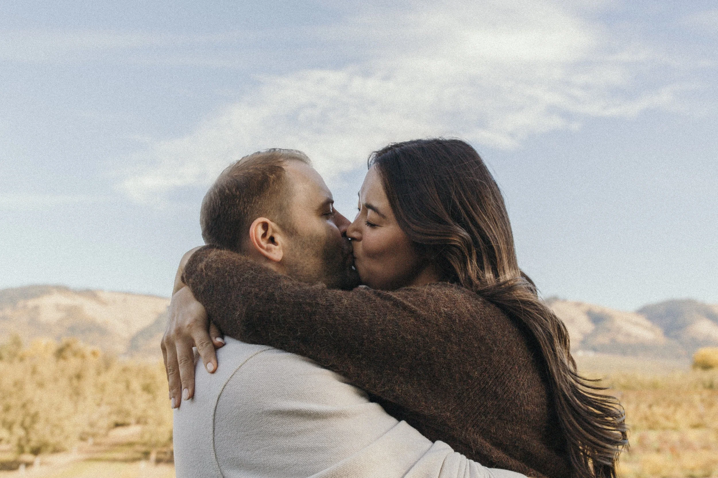 A couple kissing outdoors in a desert landscape with mountains in the background.