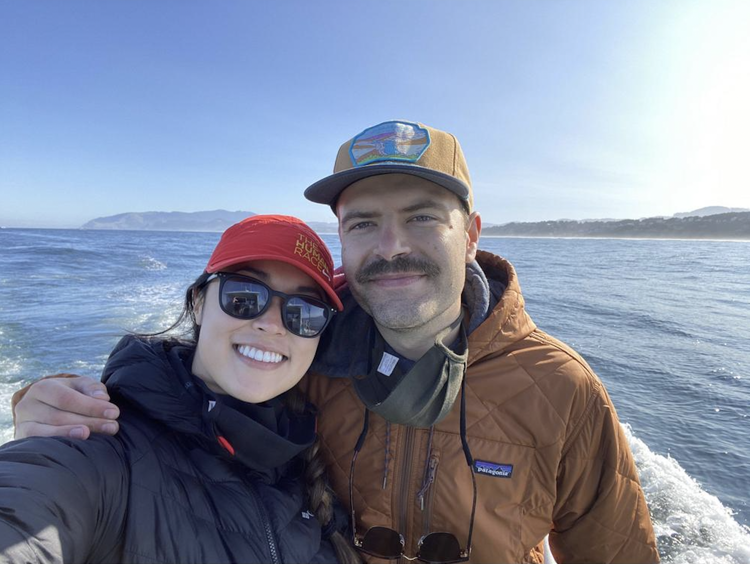 A smiling couple taking a selfie on a boat with water and distant land in the background, wearing outdoor jackets and hats.