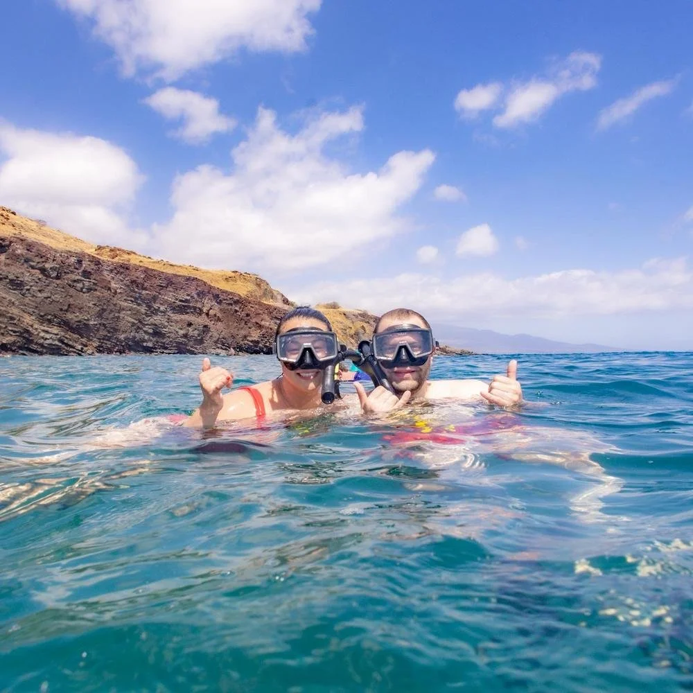 Two people wearing snorkeling masks swimming in the water near rocky shoreline, giving thumbs up and smiling.