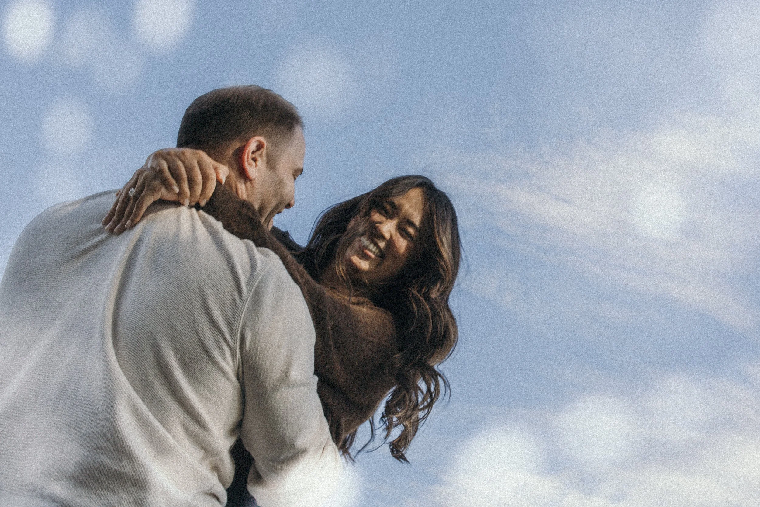A man giving a piggyback ride to a woman, both laughing against a blue sky with some clouds.