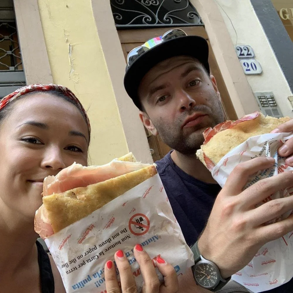 Two people holding large sandwiches wrapped in paper, standing outdoors near a building with the numbers 22 and 20 visible on the wall.