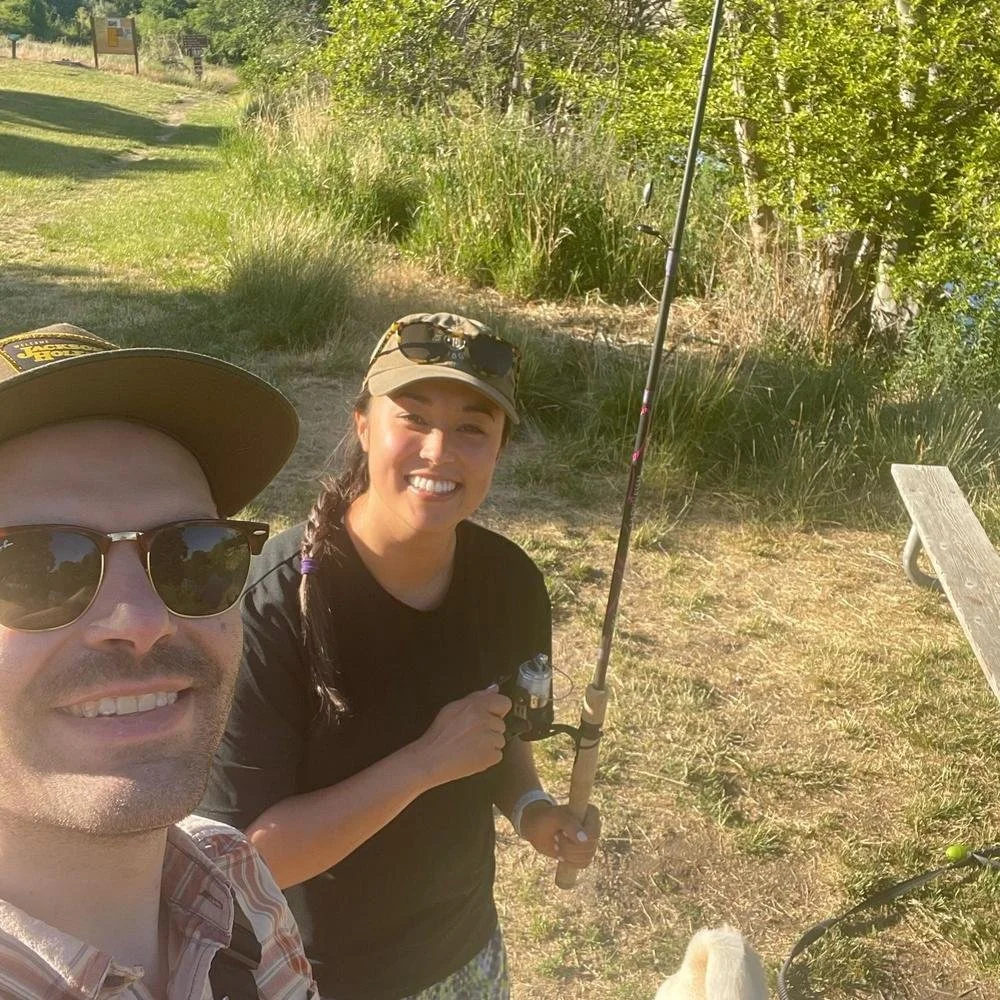 A man and a woman taking a selfie outdoors on a sunny day, with the woman holding a fishing rod. The man is wearing sunglasses and a hat, while the woman is smiling and also wearing a hat with sunglasses on top. There is a grassy area with trees in t