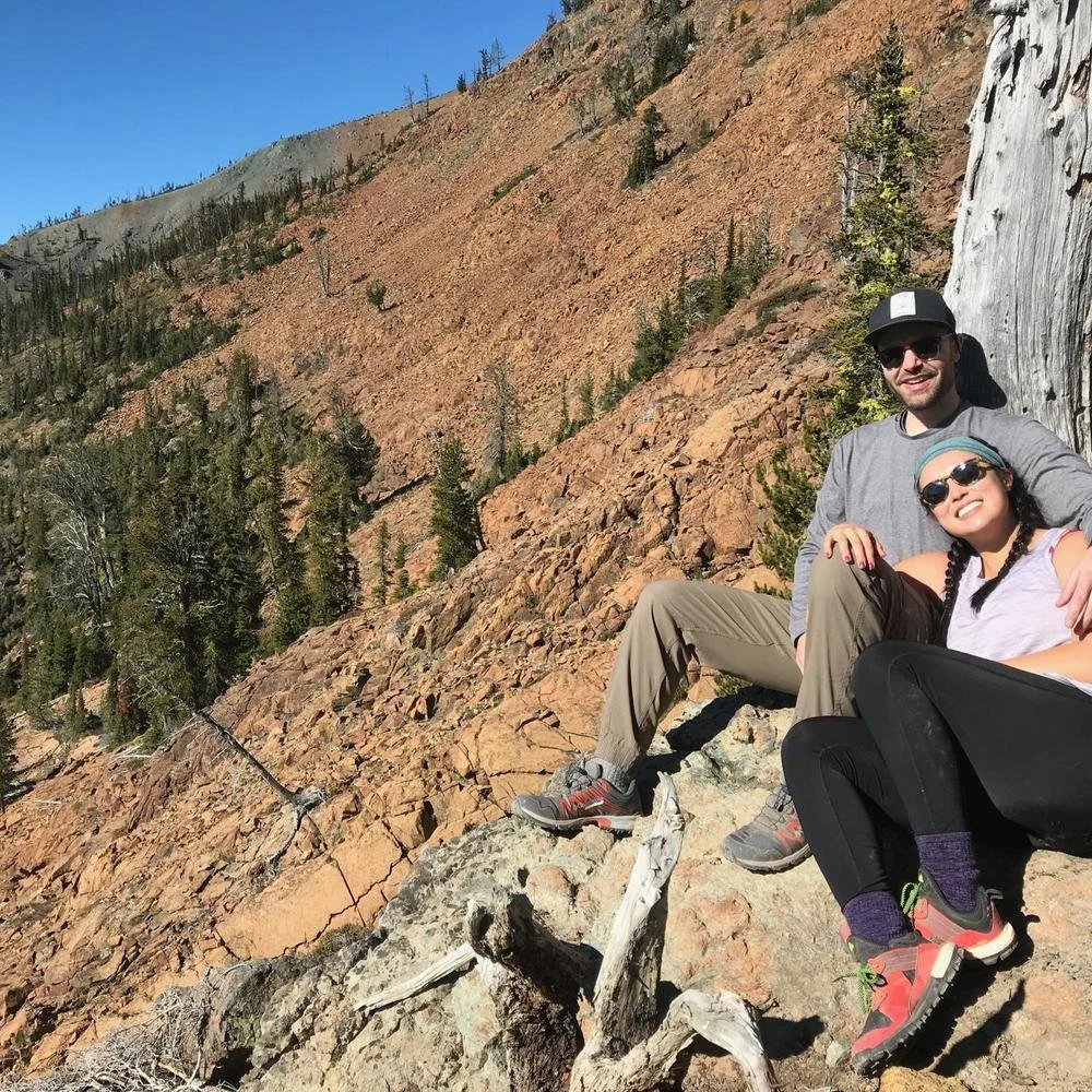 A man and woman sitting on rocky terrain in a mountainous area with trees and steep slopes, smiling at the camera on a sunny day.