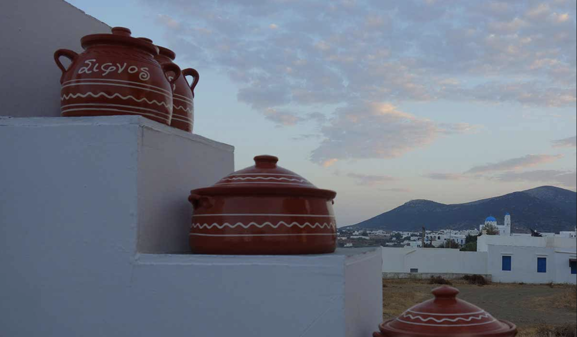 Three traditional Greek terracotta pots with white decorative patterns on a white wall ledge, with a village and mountains in the background under a partly cloudy sky.