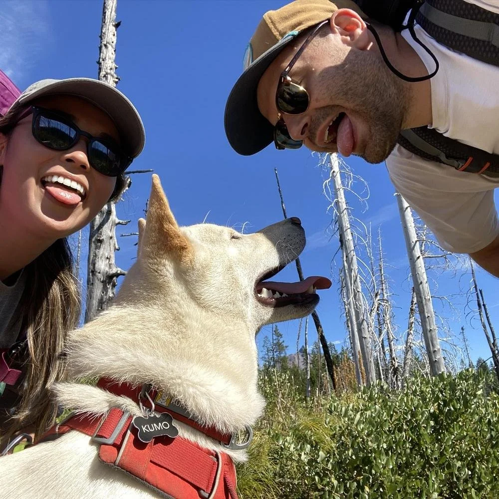 A woman and man smiling with a white dog outdoors, with a background of tall, leafless trees and a clear blue sky.