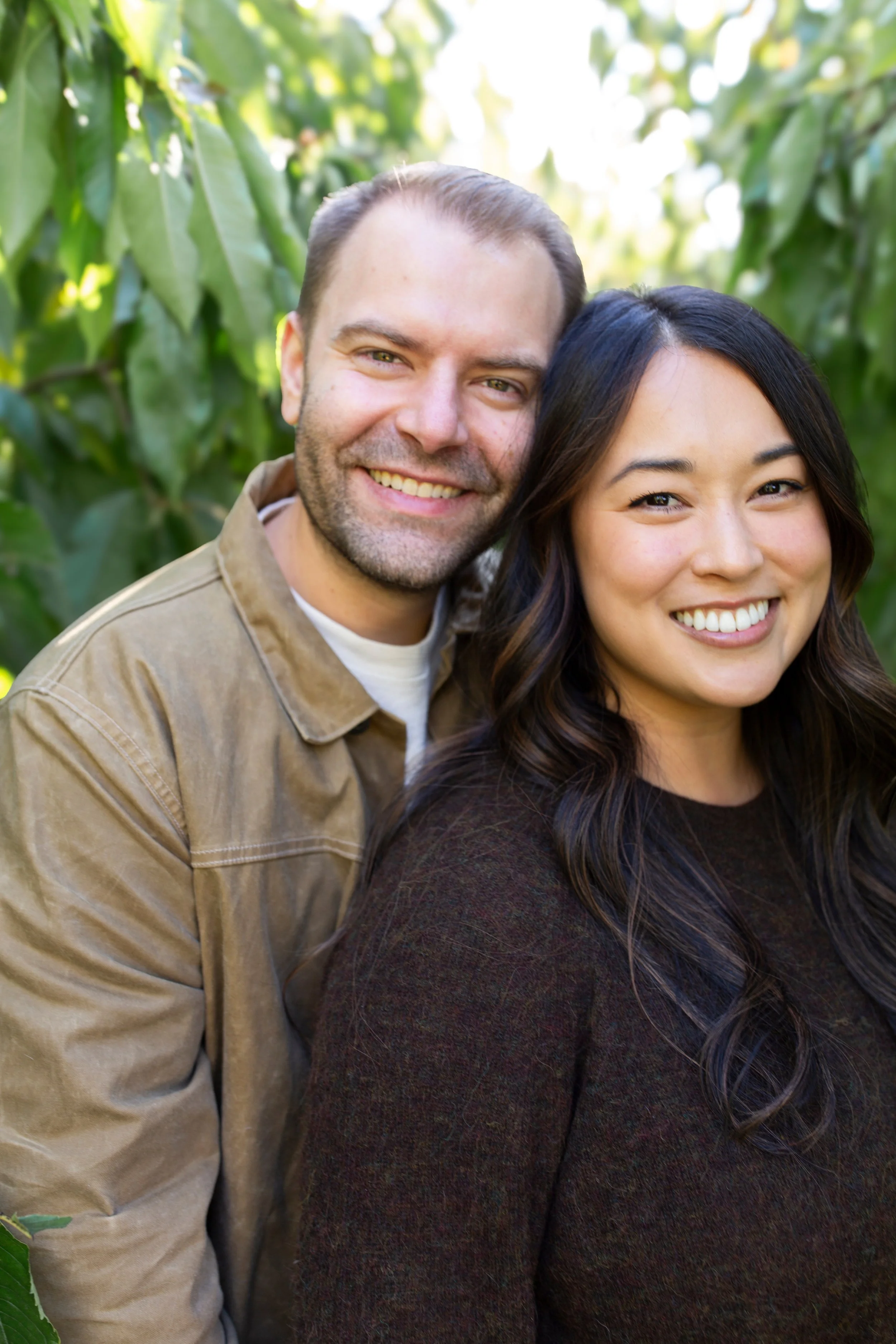 A smiling man and woman taking a selfie together in a green, leafy outdoor setting.