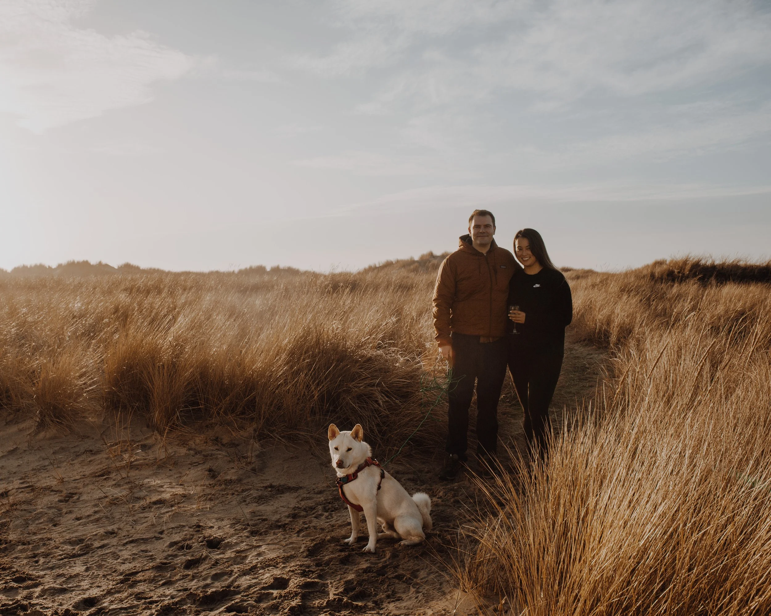 A couple standing on a sandy path in a field of tall golden grass, with a dog sitting on the ground nearby, under a blue sky with wispy clouds.