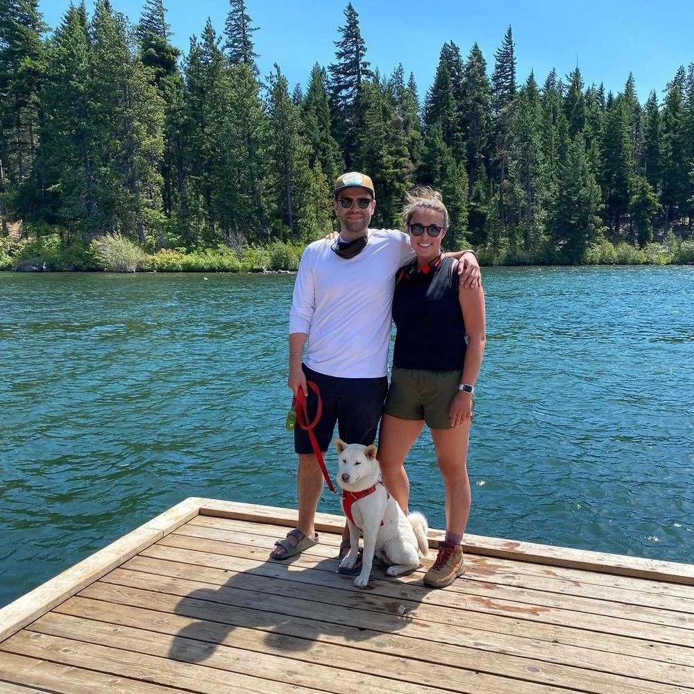 A man and woman standing on a wooden dock by a lake with a dog between them, surrounded by tall pine trees and clear blue sky.