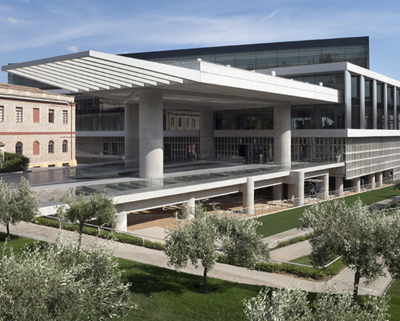 Modern architectural building with glass windows, large overhangs, and parking area underneath, surrounded by a landscaped garden with trees and pathways.