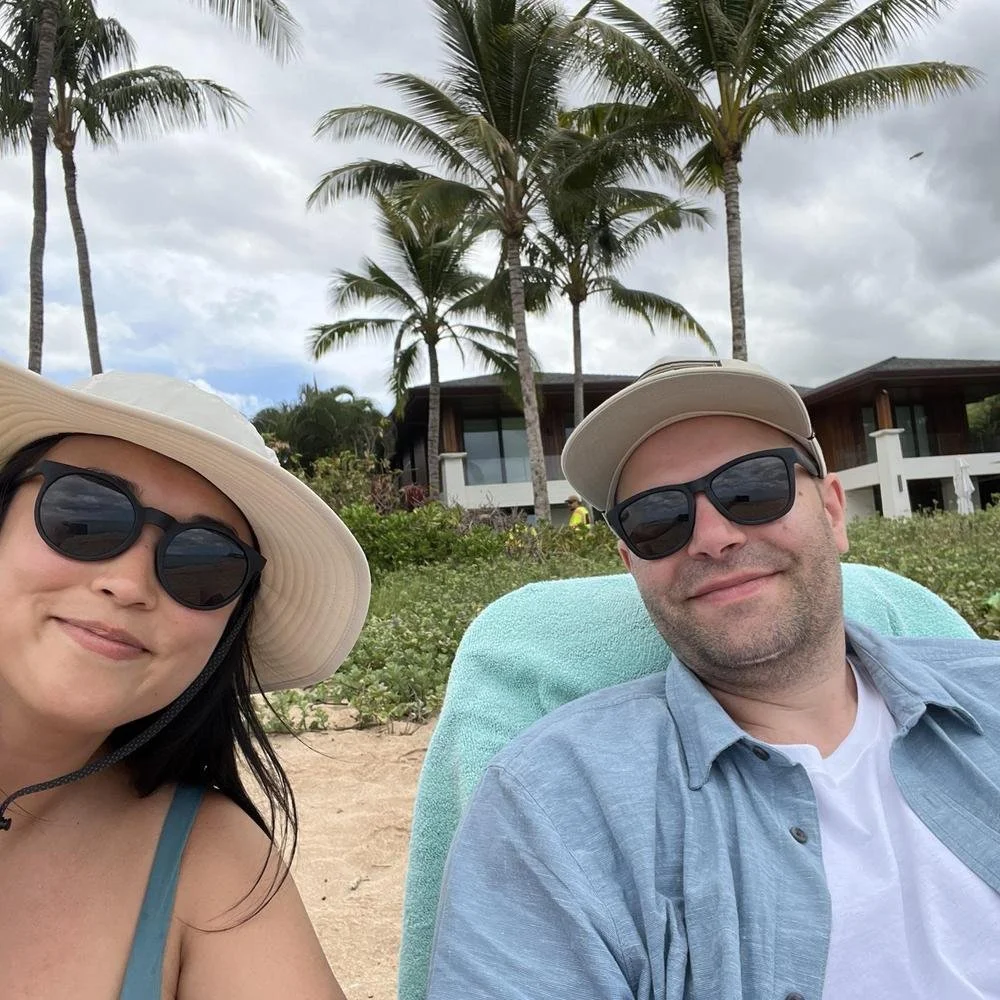 A woman and a man wearing sunglasses and hats, sitting on a beach with palm trees, modern houses, and a cloudy sky in the background.