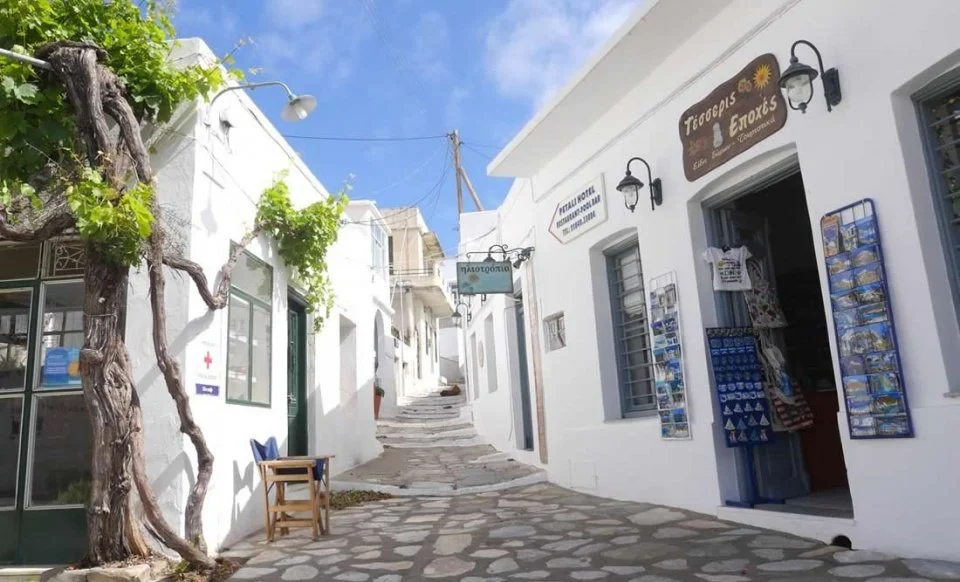 Narrow cobblestone street lined with white buildings, some with souvenirs displays outside, under a blue sky with clouds.