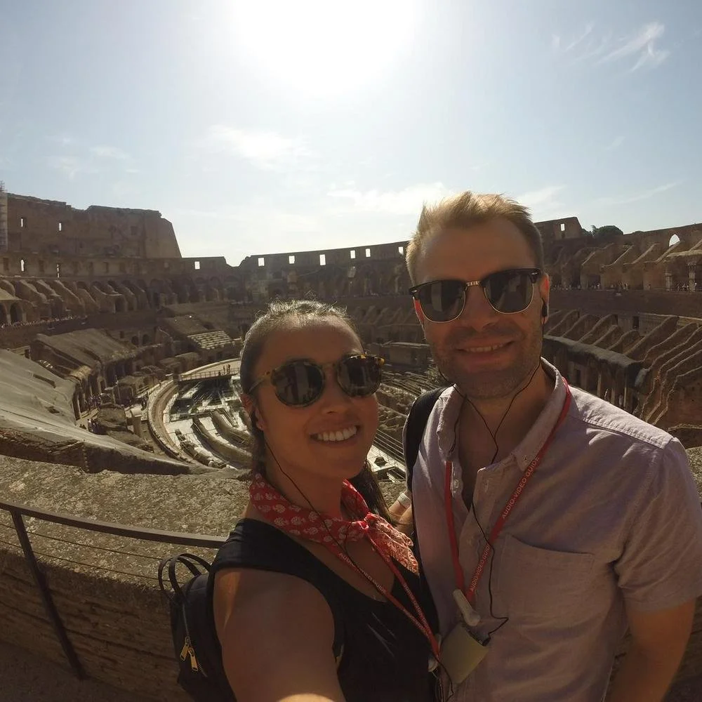 Two people taking a selfie at the ancient Roman Colosseum in Rome, Italy, with the amphitheater's interior and partial exterior visible in the background, under a bright, partly cloudy sky.