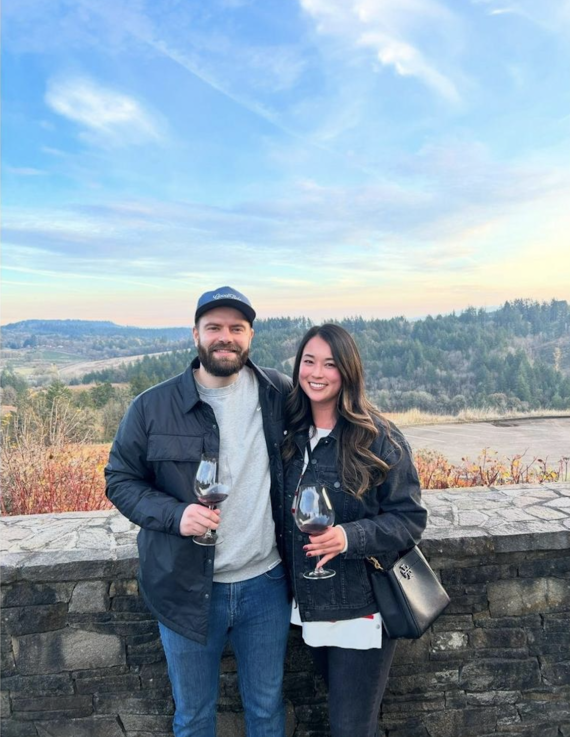 A man and woman standing outdoors near a stone wall, holding glasses of red wine, with a scenic landscape of hills and trees under a partly cloudy sky in the background.