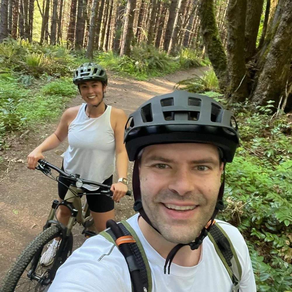 A man and woman wearing helmets riding mountain bikes on a forest trail surrounded by green foliage.