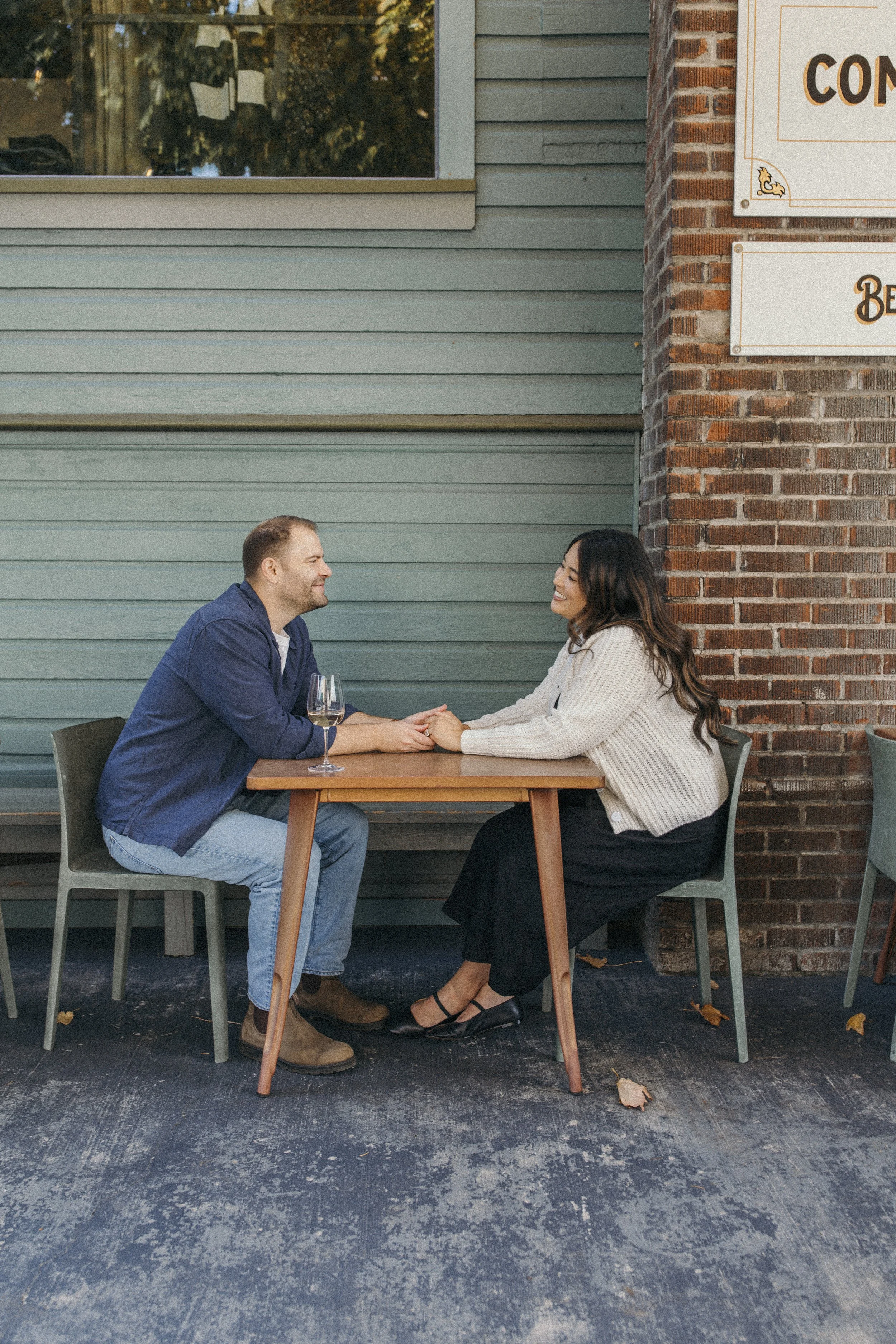 A man and woman sitting at a small outdoor table holding hands and smiling at each other, with a glass of wine on the table, in front of a building with blue siding and brick wall.