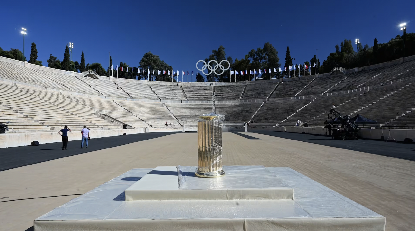 Empty outdoor amphitheater with the Olympic rings overhead, flags around the top, and a stage with a trophy in the center, under a clear and sunny sky.